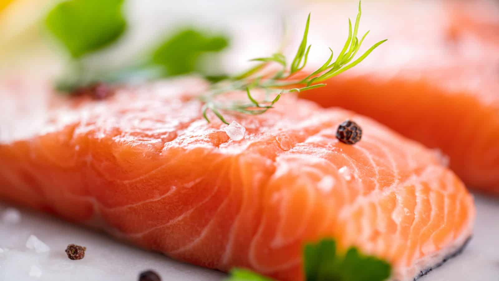 Close-up of a raw salmon fillet garnished with a sprig of dill, whole black peppercorns, coarse salt, and fresh parsley leaves.