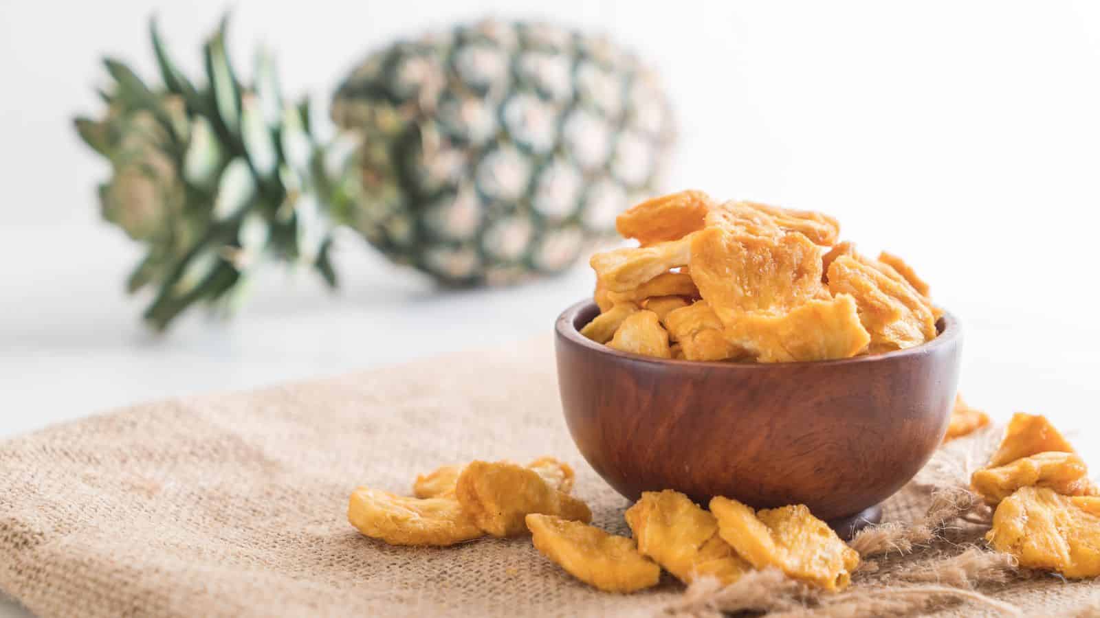 A wooden bowl filled with dried pineapple pieces sits on a burlap cloth, with a whole pineapple placed in the background on a white surface.