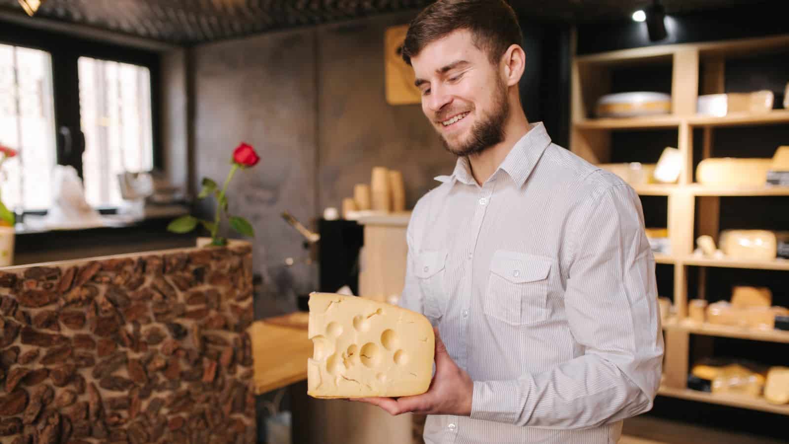 A man in a white shirt stands indoors, smiling and holding a large block of Swiss cheese with holes. Shelves with more cheese and a single red rose in a vase are visible in the background.