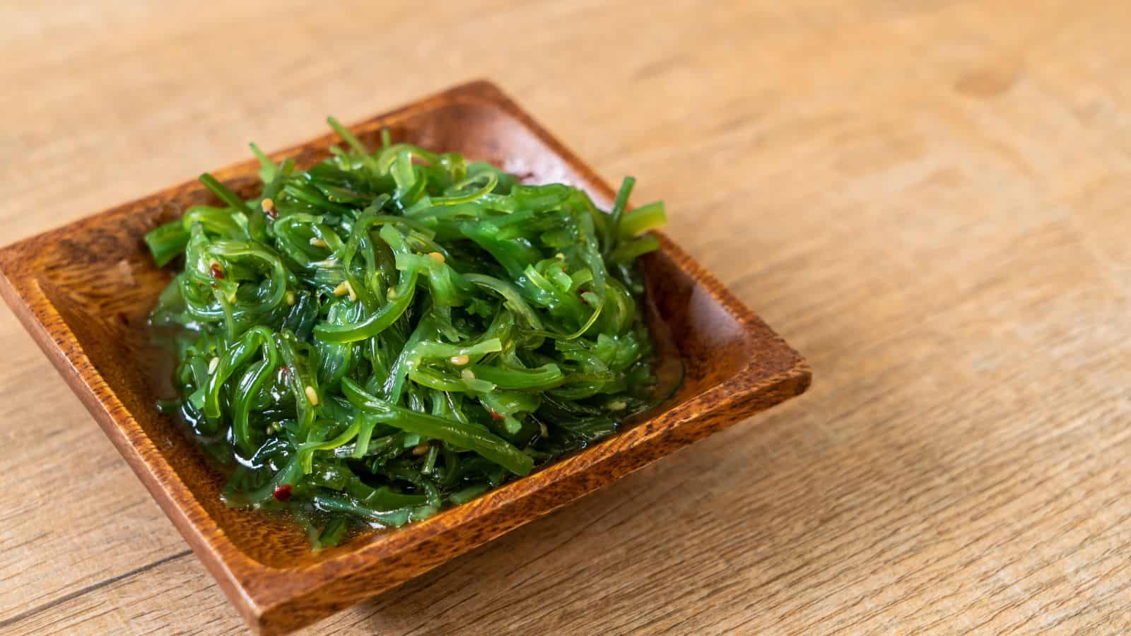 A small wooden bowl filled with green seaweed salad, placed on a light wooden surface. The seaweed is glossy and garnished with sesame seeds and red pepper flakes.