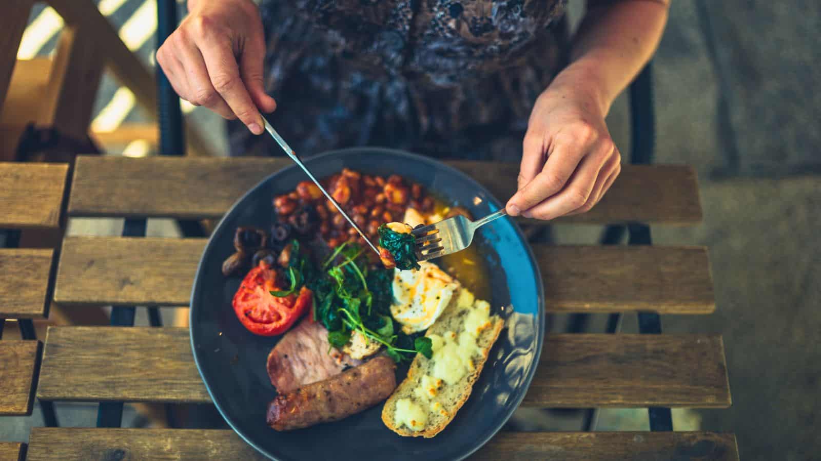 A person sits at a wooden table, holding a fork and knife over a plate with grilled tomato, sausage, beans, greens, eggs, olives, and a baked potato. The person is about to eat.