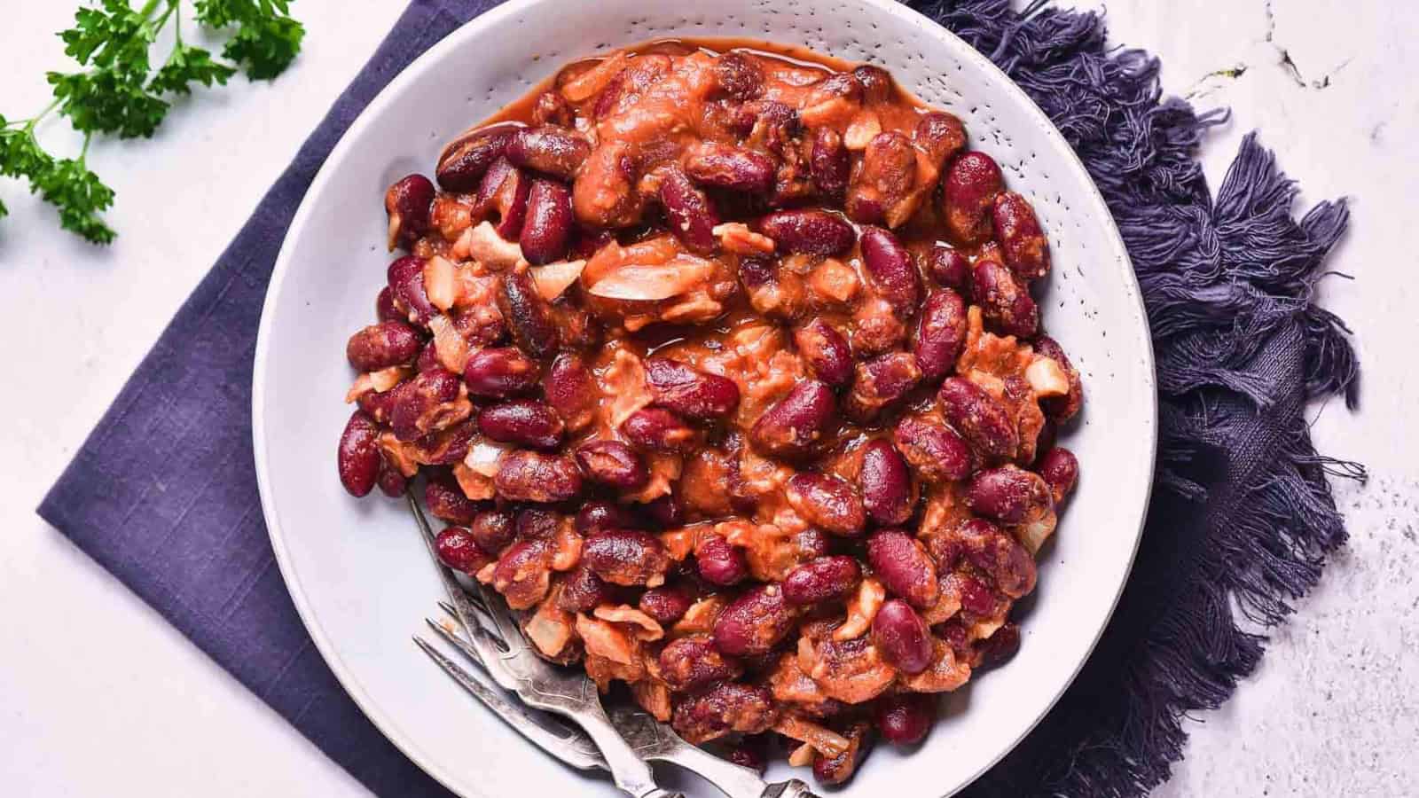 A white bowl filled with a red kidney bean stew, including visible pieces of meat or vegetables, sits on a dark cloth napkin with a fork and spoon beside it. A small bunch of parsley is on the table nearby.