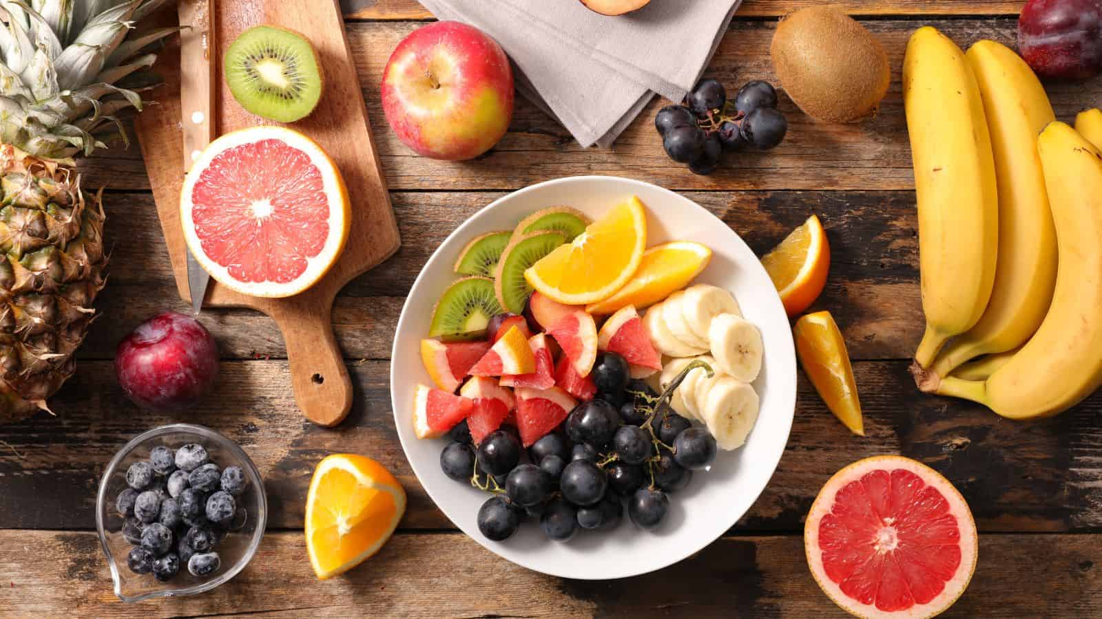 A white plate filled with sliced grapefruit, kiwi, banana, orange, and black grapes sits on a rustic wooden table surrounded by whole and cut fruit, including bananas, apple, pineapple, plums, blueberries, kiwi, and grapefruit.