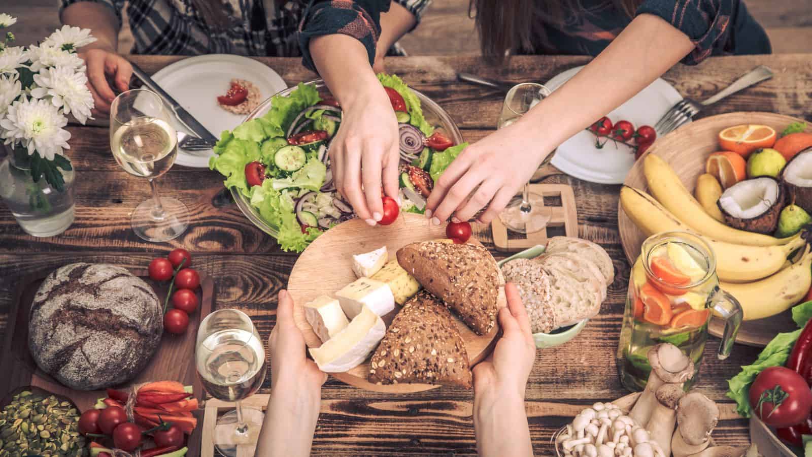 A wooden table set with various foods, including bread, cheese, salad, fruits, and wine. Three people are reaching for food, and flowers are placed in a vase on the table.