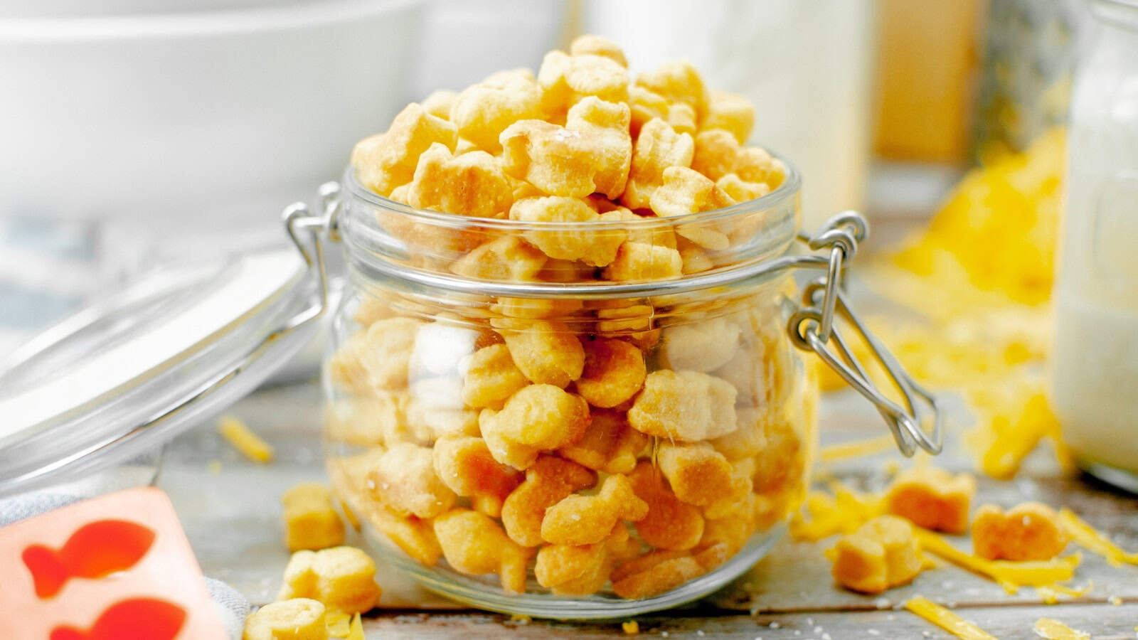 A glass jar filled with fish-shaped cheese crackers on a wooden table, surrounded by scattered crackers and cheese shreds.