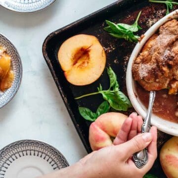 A hand serves peach cobbler from a white baking dish onto a plate. The dish sits on a tray with fresh peaches, peach halves, and basil leaves nearby. Two bowls of cobbler are also on the table.