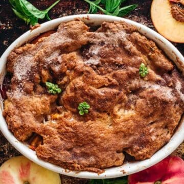 A baked peach cobbler in an oval dish, topped with brown crust and garnished with small green herbs, surrounded by whole and halved peaches and fresh basil leaves on a dark surface.