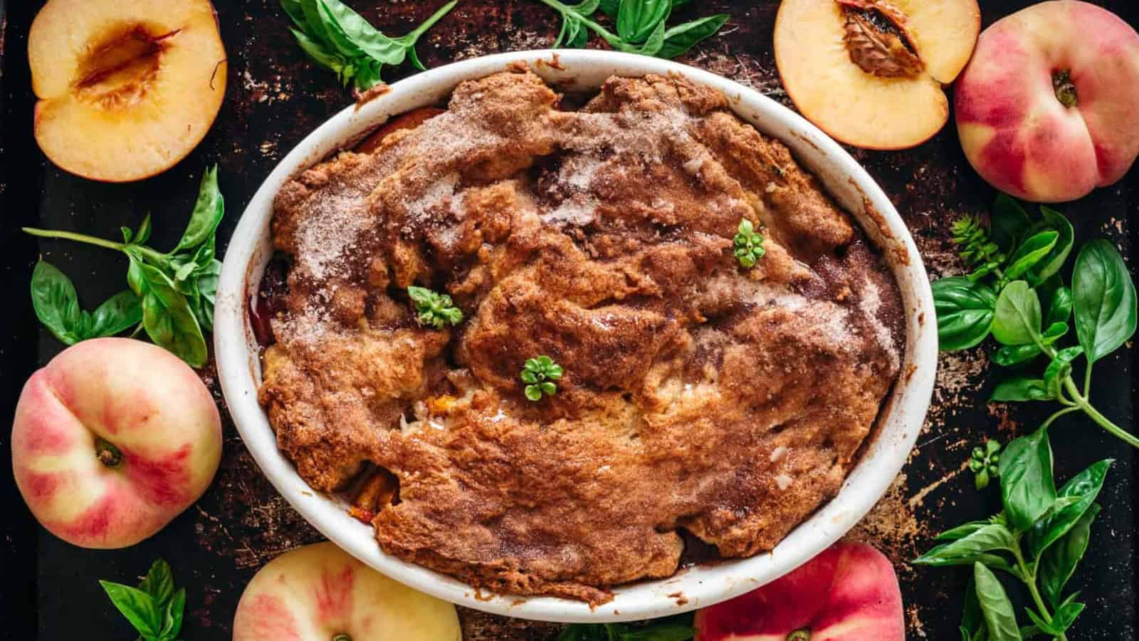 A baked peach cobbler in a white oval dish, topped with sprigs of fresh herbs, is surrounded by whole and halved peaches and green basil leaves on a dark surface.
