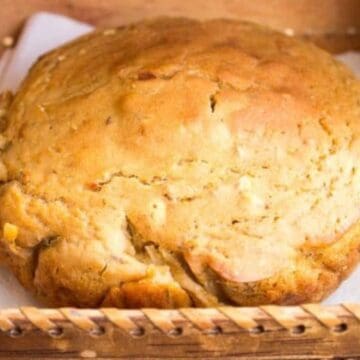 A round, golden-brown loaf of bread with a slightly cracked surface sits on a white napkin inside a wooden tray.