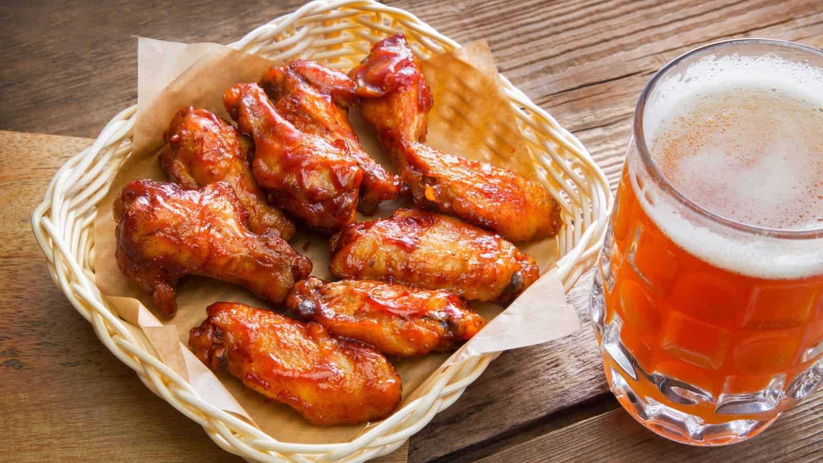 A basket of saucy chicken wings sits on parchment paper next to a glass of beer on a wooden surface.