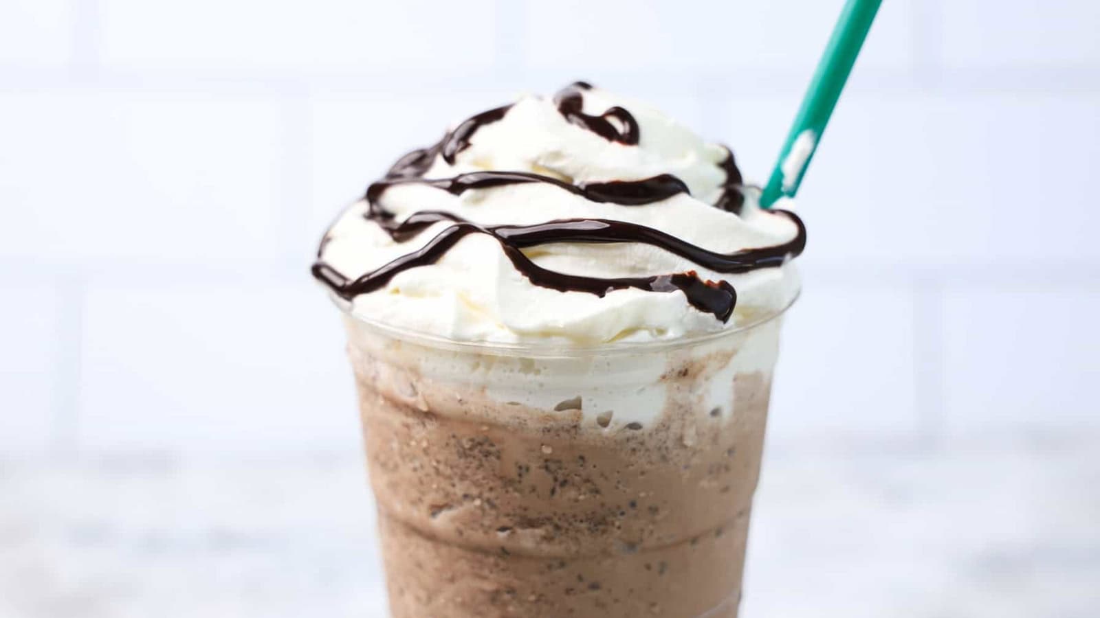 A blended iced mocha drink topped with whipped cream and chocolate syrup, served in a clear plastic cup with a green straw against a light background.