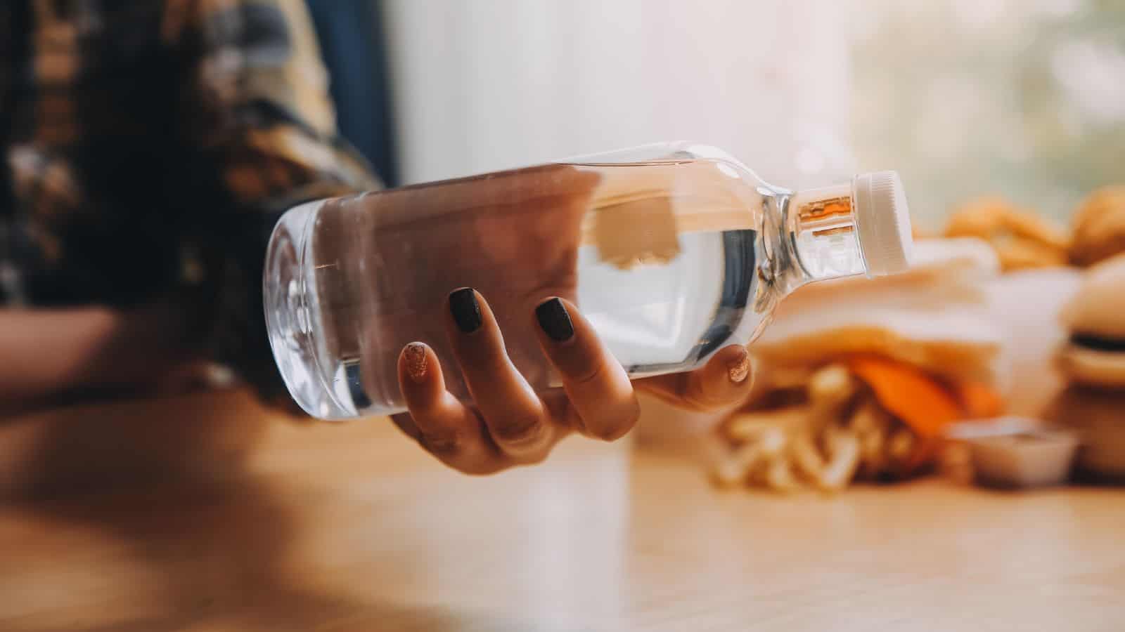 A person holding a clear plastic bottle filled with water over a wooden table, with fast food items like fries and burgers blurred in the background.