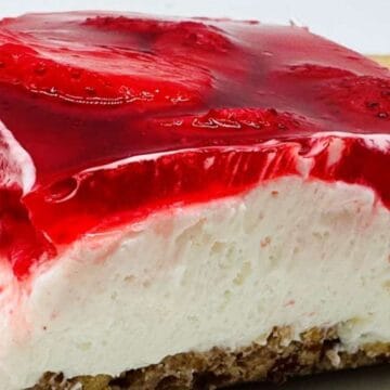 A close-up of a slice of layered dessert with a crumbly brown base, thick white cream middle layer, and a glossy red strawberry gelatin topping with visible strawberry slices.