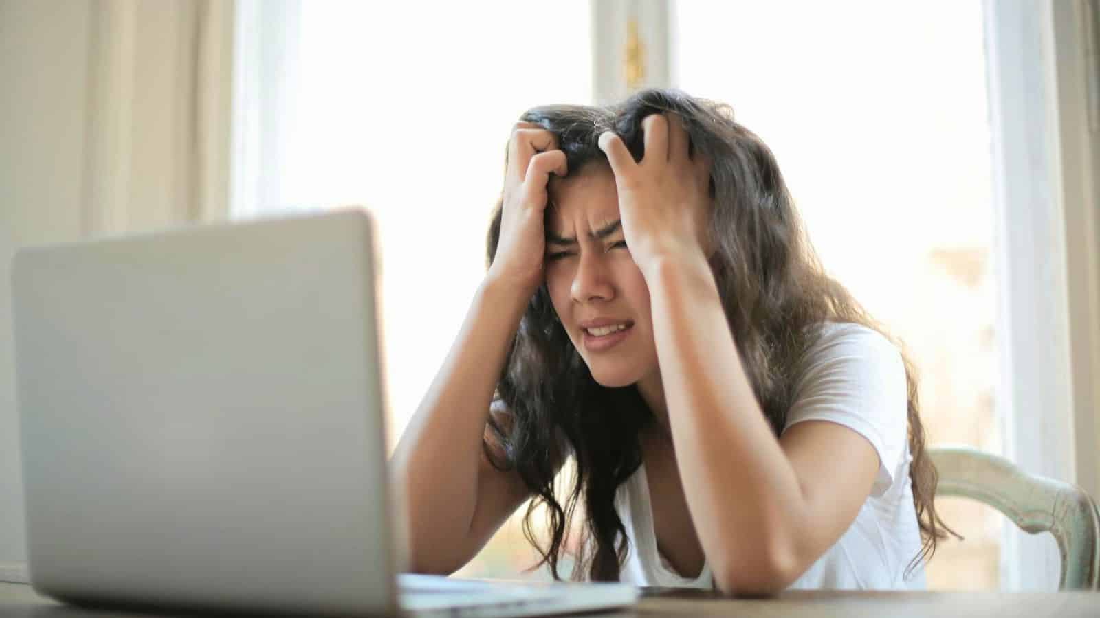 A young woman sits at a table in front of an open laptop, holding her head with both hands and appearing frustrated or stressed. She is wearing a white shirt, and the room is brightly lit by natural light.