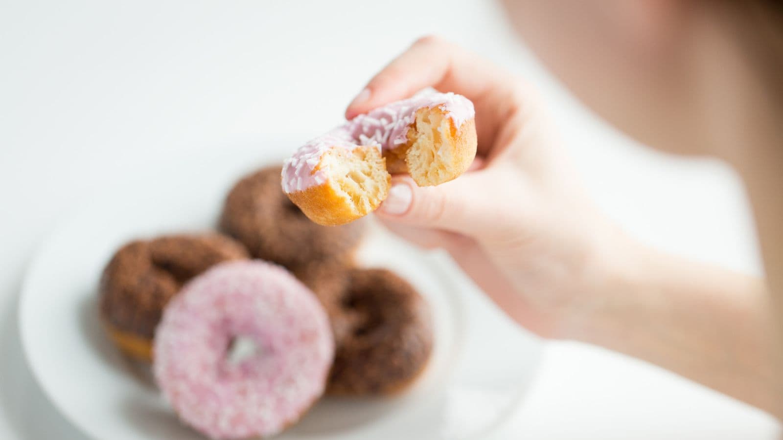 A person holds a halved pink-frosted donut, showing its inside. In the background, a white plate contains more donuts with chocolate and pink frosting.