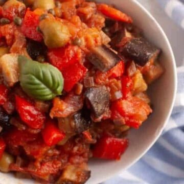 A bowl of caponata with diced eggplant, red bell peppers, tomatoes, and a basil leaf garnish, placed beside a blue and white striped cloth.