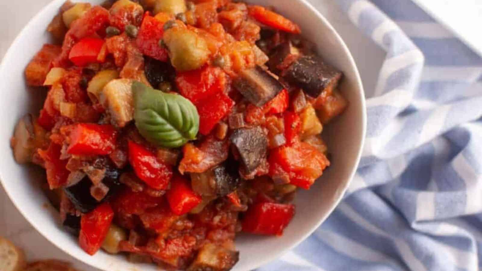 A bowl of caponata with diced eggplant, red bell peppers, tomatoes, and a basil leaf garnish, placed beside a blue and white striped cloth.