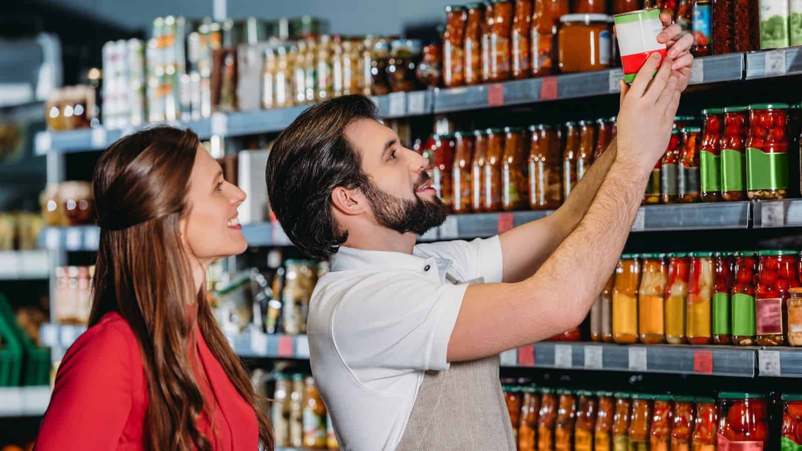 A man in a light shirt and apron reaches for a jar on a grocery store shelf while a woman in a red top stands beside him. Shelves are filled with jars of preserved food.