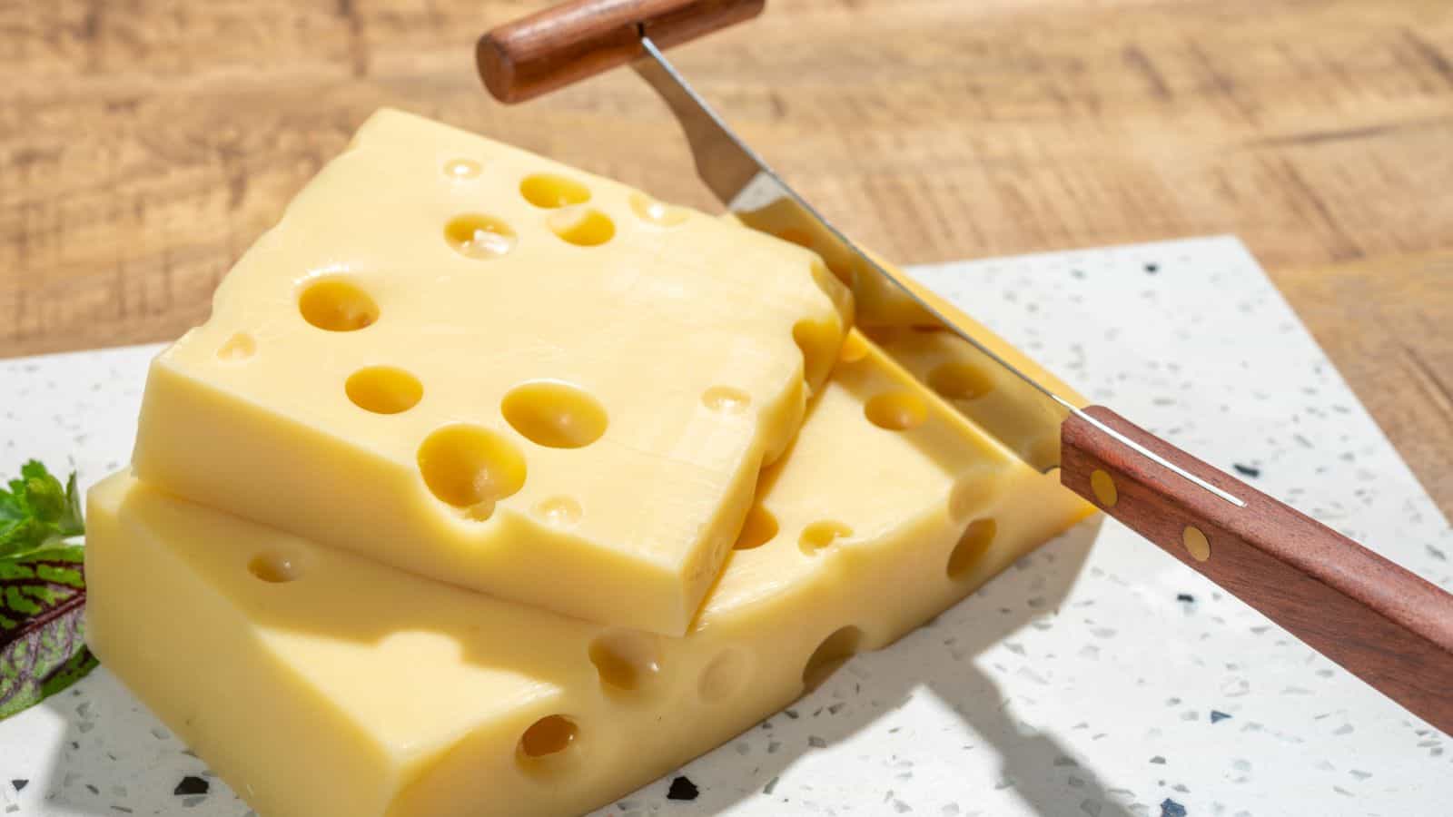 Two slices of Swiss cheese with holes are stacked on a white speckled cutting board. A cheese slicer with a wooden handle rests on top. The background is a wooden surface.