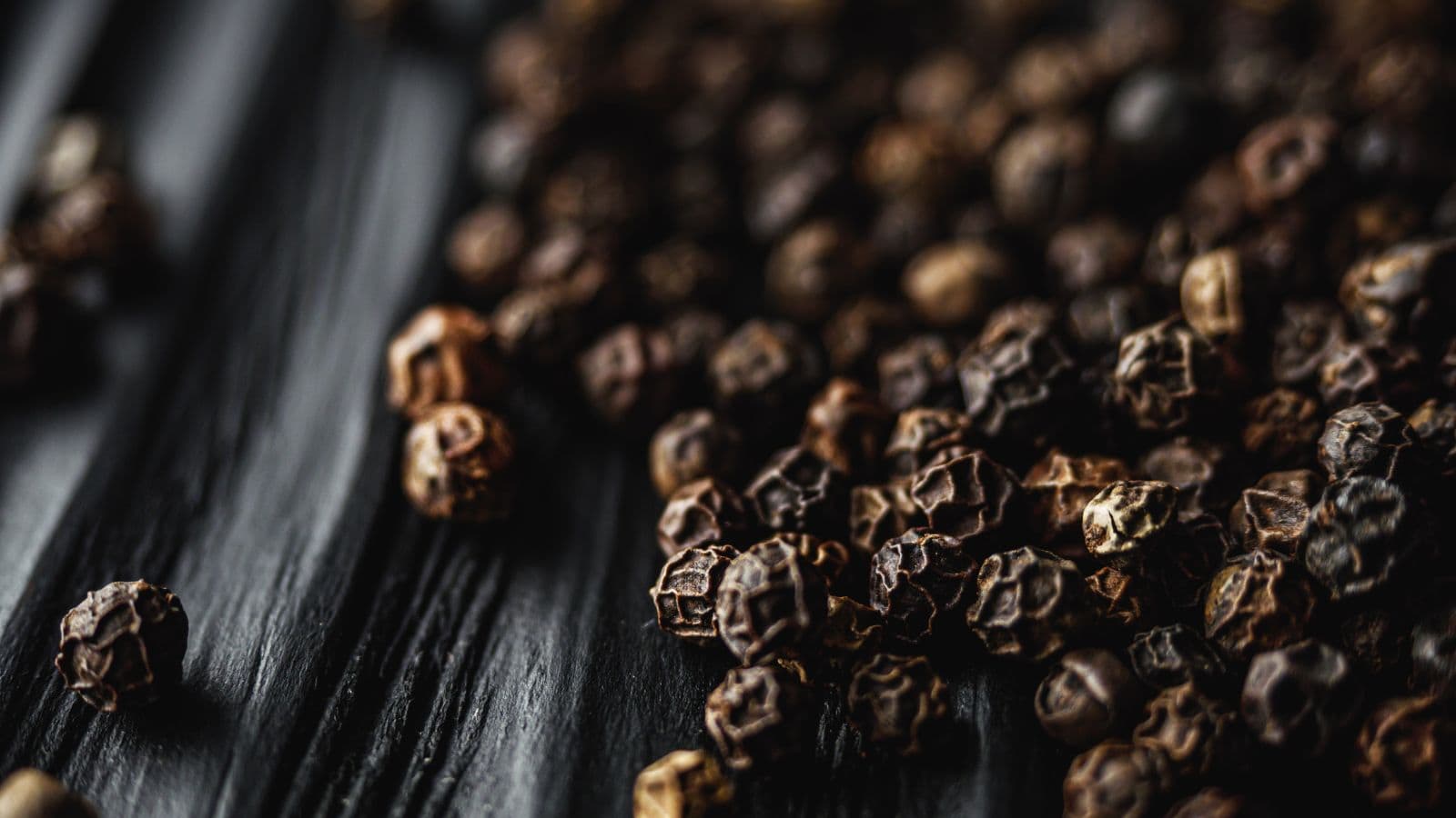 Close-up view of dried black peppercorns scattered on a dark textured surface, with some in sharp focus and others blurred in the background.