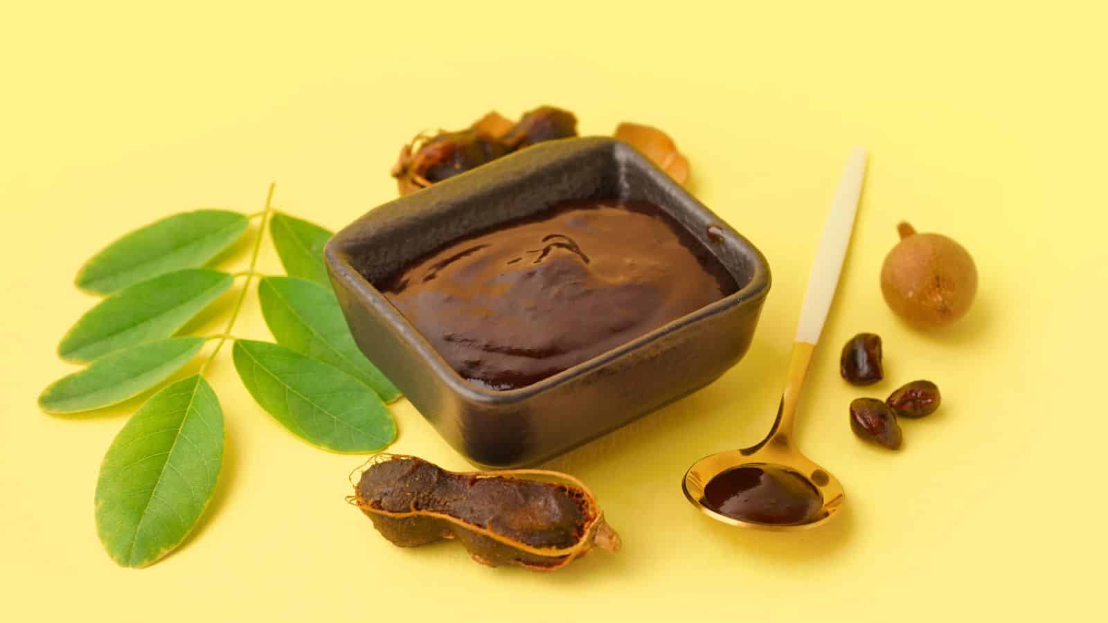 A small square bowl filled with thick brown tamarind paste is surrounded by tamarind pods, seeds, a sprig of green leaves, and a golden spoon with paste on a yellow background.