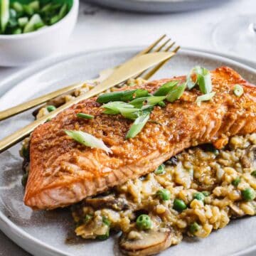 A cooked salmon fillet sits on top of a bed of risotto with peas and mushrooms, garnished with sliced green onions, on a round plate with a fork and knife. A bowl of green onions is in the background.