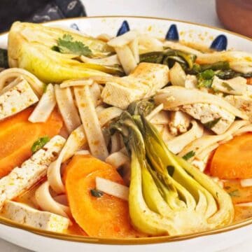 A bowl of soup with sliced tofu, baby bok choy, carrot rounds, and wide rice noodles in a light orange broth, garnished with green herbs.