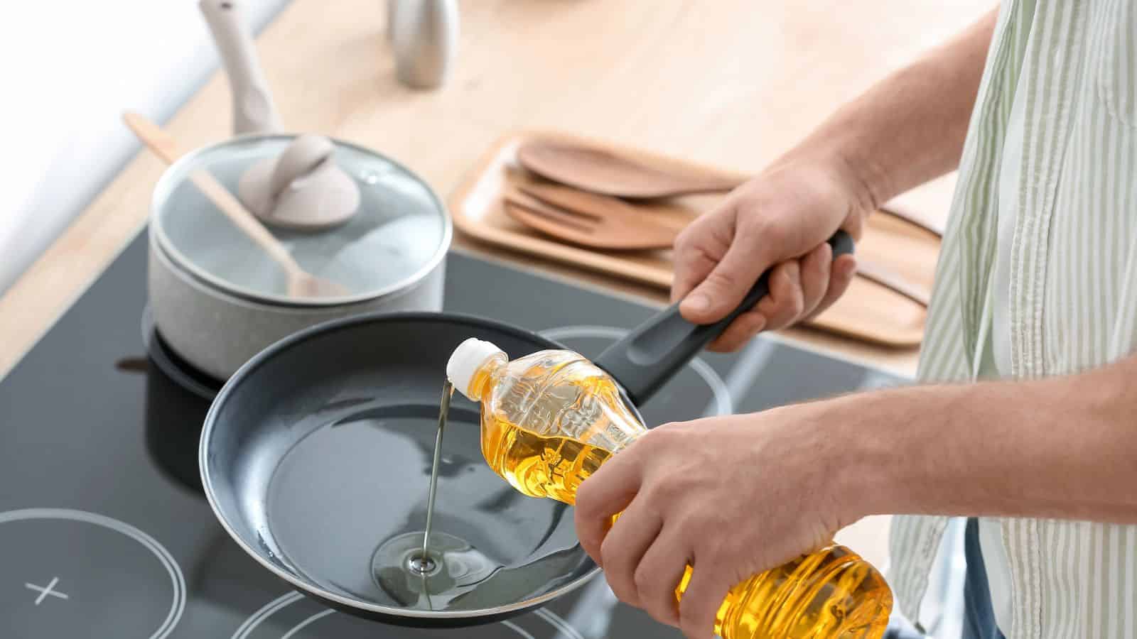 A person pours cooking oil from a plastic bottle into a black frying pan on a stovetop. A pot with a lid, wooden utensils, and a tray are visible on the countertop in the background.