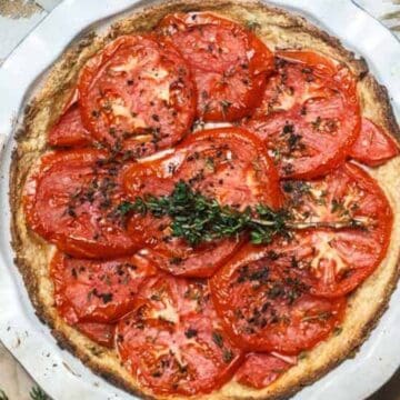 A baked tomato tart topped with sliced tomatoes and fresh herbs, in a metal pie dish, sits on a brown crumpled paper surface with a white patterned cloth partially visible in the background.