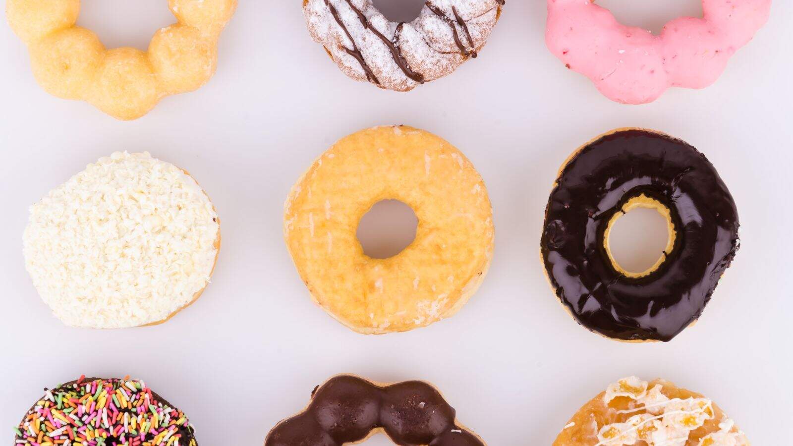 Nine assorted donuts are arranged in a three-by-three grid on a white background. The donuts have various toppings including chocolate glaze, pink icing, powdered sugar, sprinkles, and shredded coconut.