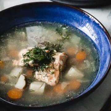 A blue bowl filled with clear soup containing pieces of fish, carrots, and potatoes, topped with fresh herbs. A silver spoon is placed on the right side of the bowl.