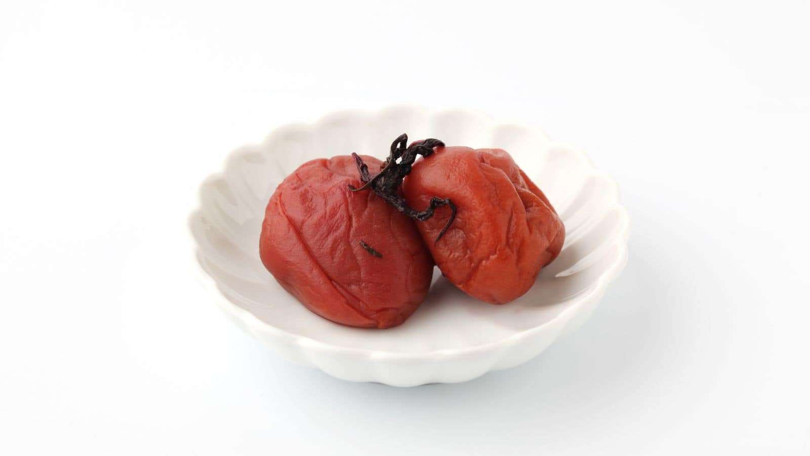 Two pickled plums, known as umeboshi, are placed on a small white scalloped dish against a plain white background.