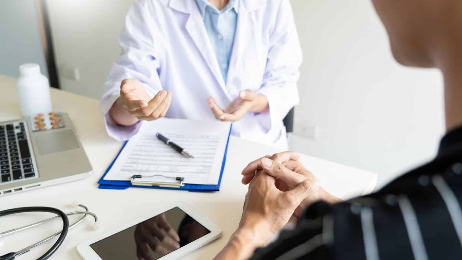 A person in a white lab coat sits at a desk talking to another person across from them. A clipboard with papers, a pen, a stethoscope, a tablet, and a laptop are on the desk.