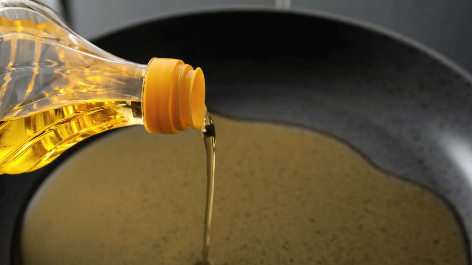 A plastic bottle with a yellow cap pours vegetable oil into a black frying pan, creating a thin layer of oil on the pan’s surface.