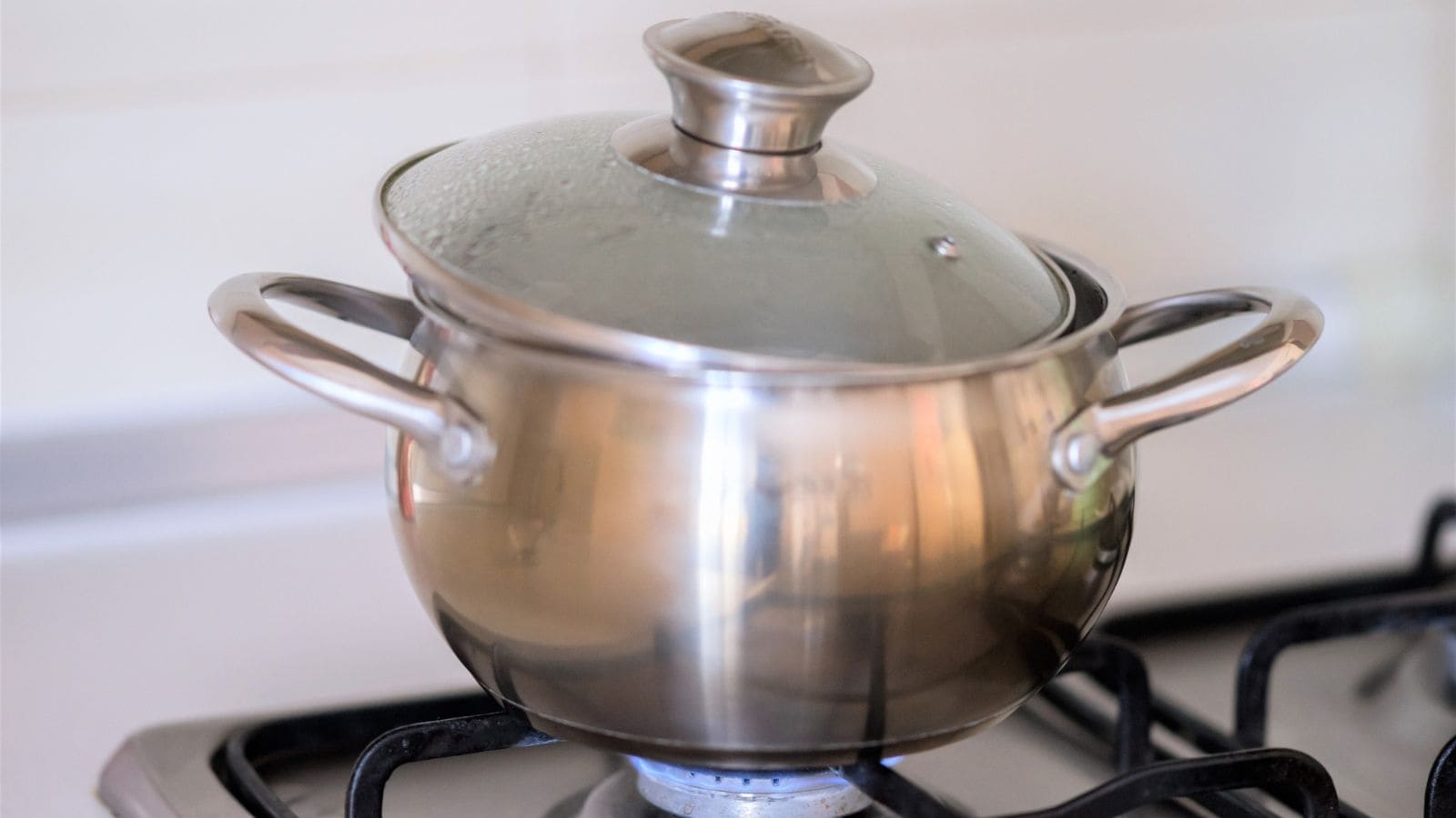 A stainless steel pot with a glass lid is sitting on a gas stove burner with the flame on. The pot appears to be in use, as condensation is visible on the lid. The background shows a countertop and wall.