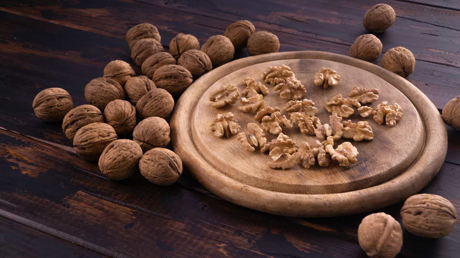Whole walnuts are scattered on a dark wooden surface next to a round wooden board with several walnut halves and pieces arranged on top.