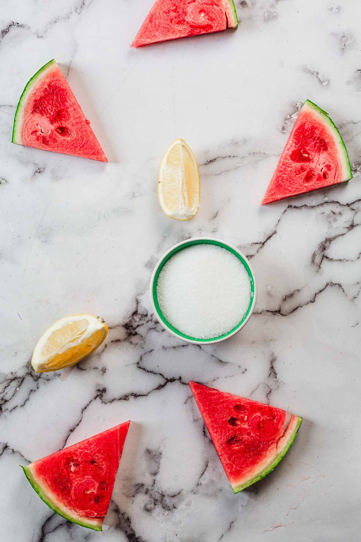Five watermelon slices, resembling Watermelon Popsicles, two lemon wedges, and a small bowl of white sugar are arranged on a white marble surface.