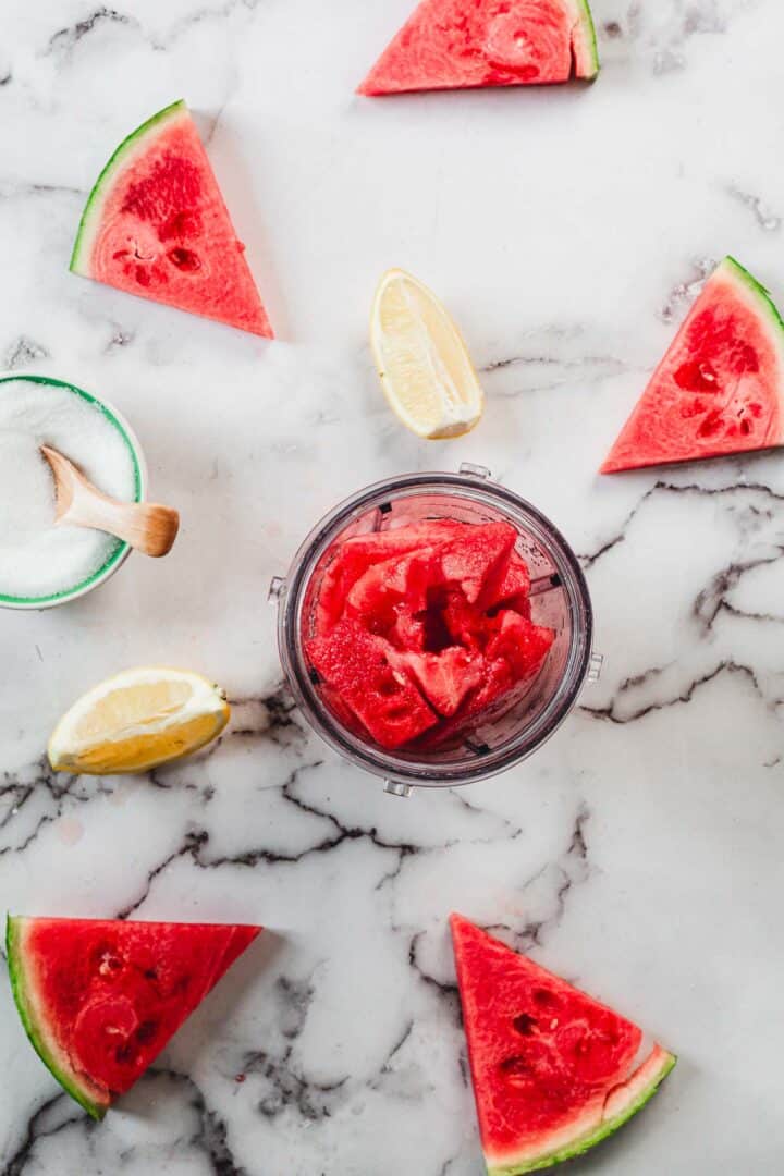 A blender cup filled with watermelon chunks sits on a marble countertop, surrounded by several watermelon slices, a bowl of sugar with a wooden spoon, two lemon wedges, and all the essentials for making refreshing Watermelon Popsicles.