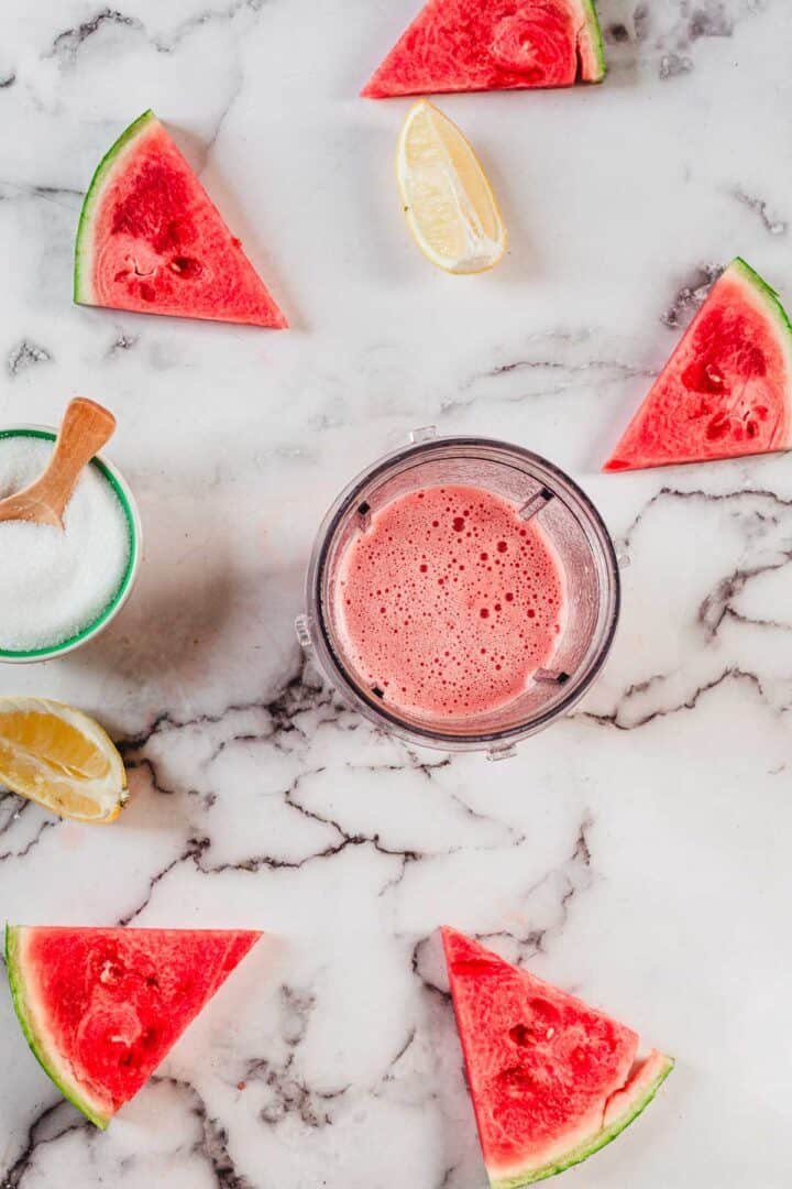 A blender cup filled with pink watermelon juice sits on a marble surface, surrounded by watermelon slices, lemon wedges, and a small bowl of sugar with a wooden spoon—perfect for making refreshing Watermelon Popsicles.