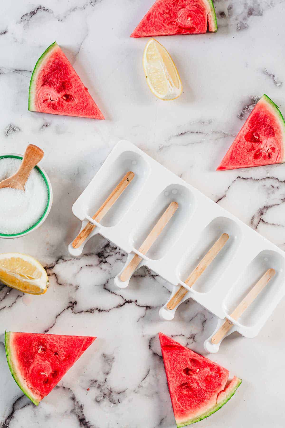 A white popsicle mold with wooden sticks sits on a marble surface surrounded by watermelon slices, lemon wedges, and a small bowl of sugar—perfect for making refreshing Watermelon Popsicles.