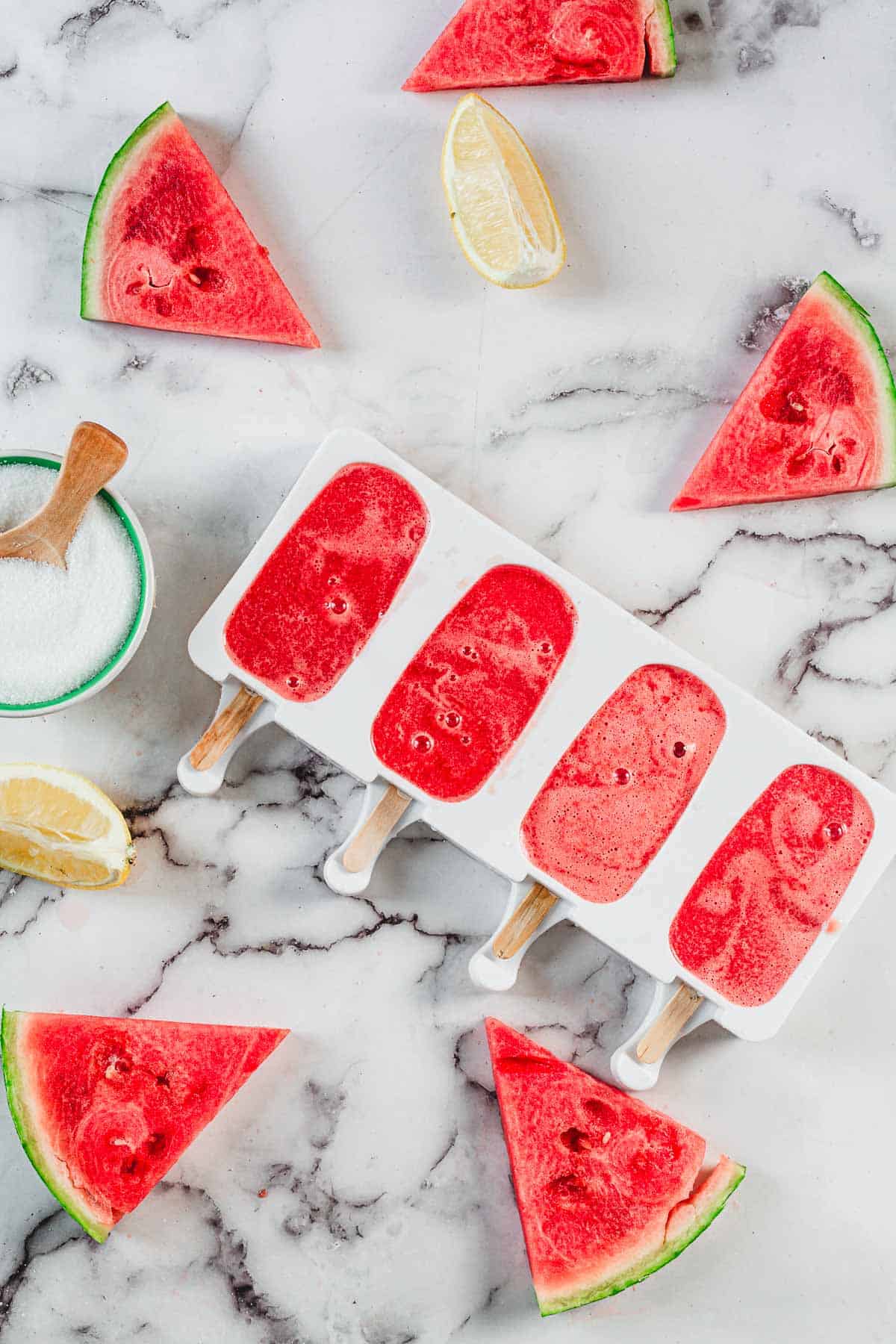 A white popsicle mold holds four refreshing Watermelon Popsicles. Surrounding the mold are fresh watermelon slices, lemon wedges, and a small bowl of sugar with a wooden spoon, all arranged on a marble surface.