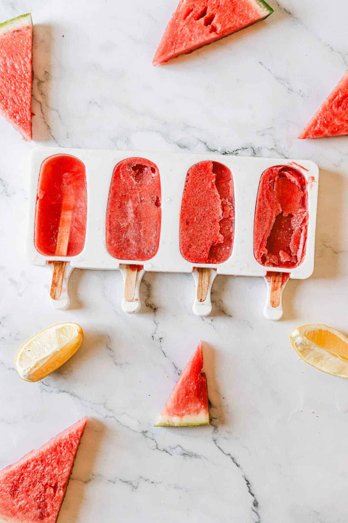 A white popsicle mold holds four refreshing Watermelon Popsicles on wooden sticks, with watermelon slices and lemon wedges arranged on a white marble surface around the mold.