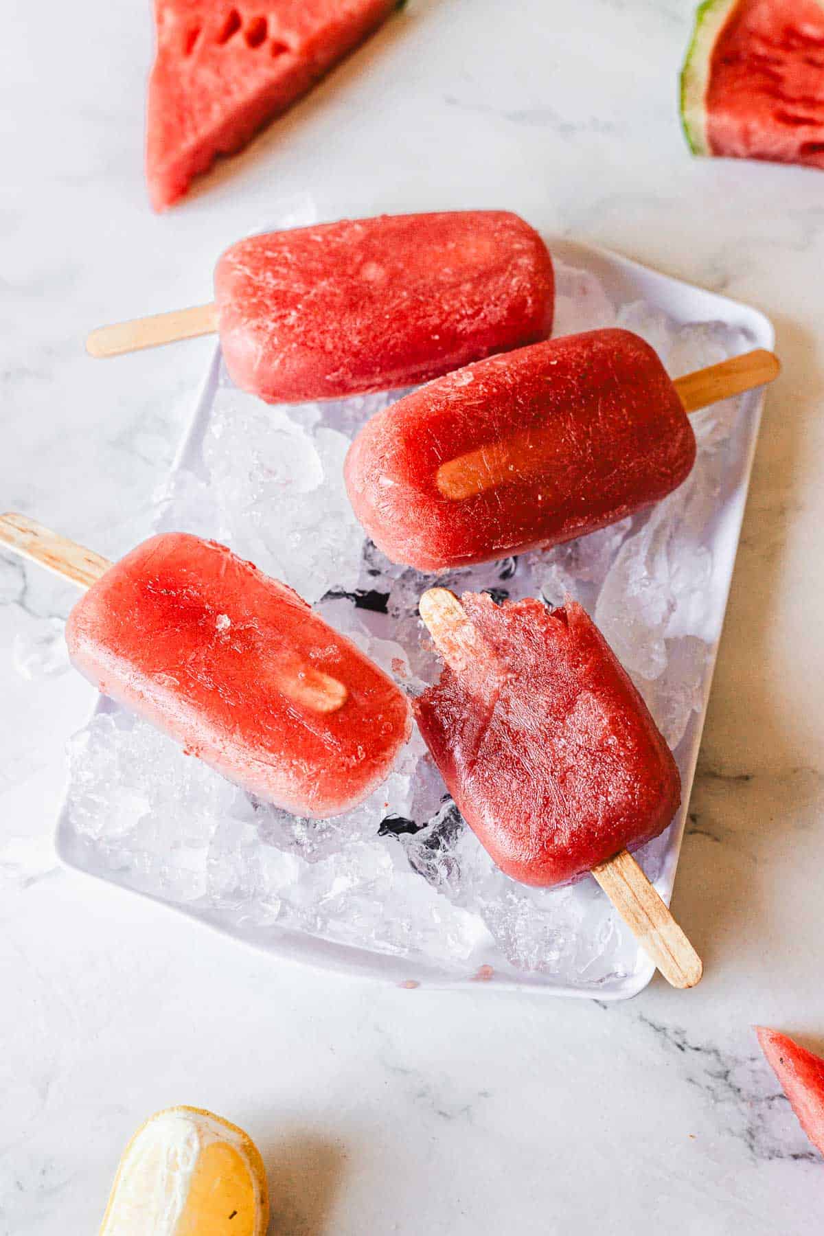 Four refreshing Watermelon Popsicles on wooden sticks rest on a tray filled with ice cubes. Slices of watermelon and a lemon wedge are partially visible around the tray on a white surface.