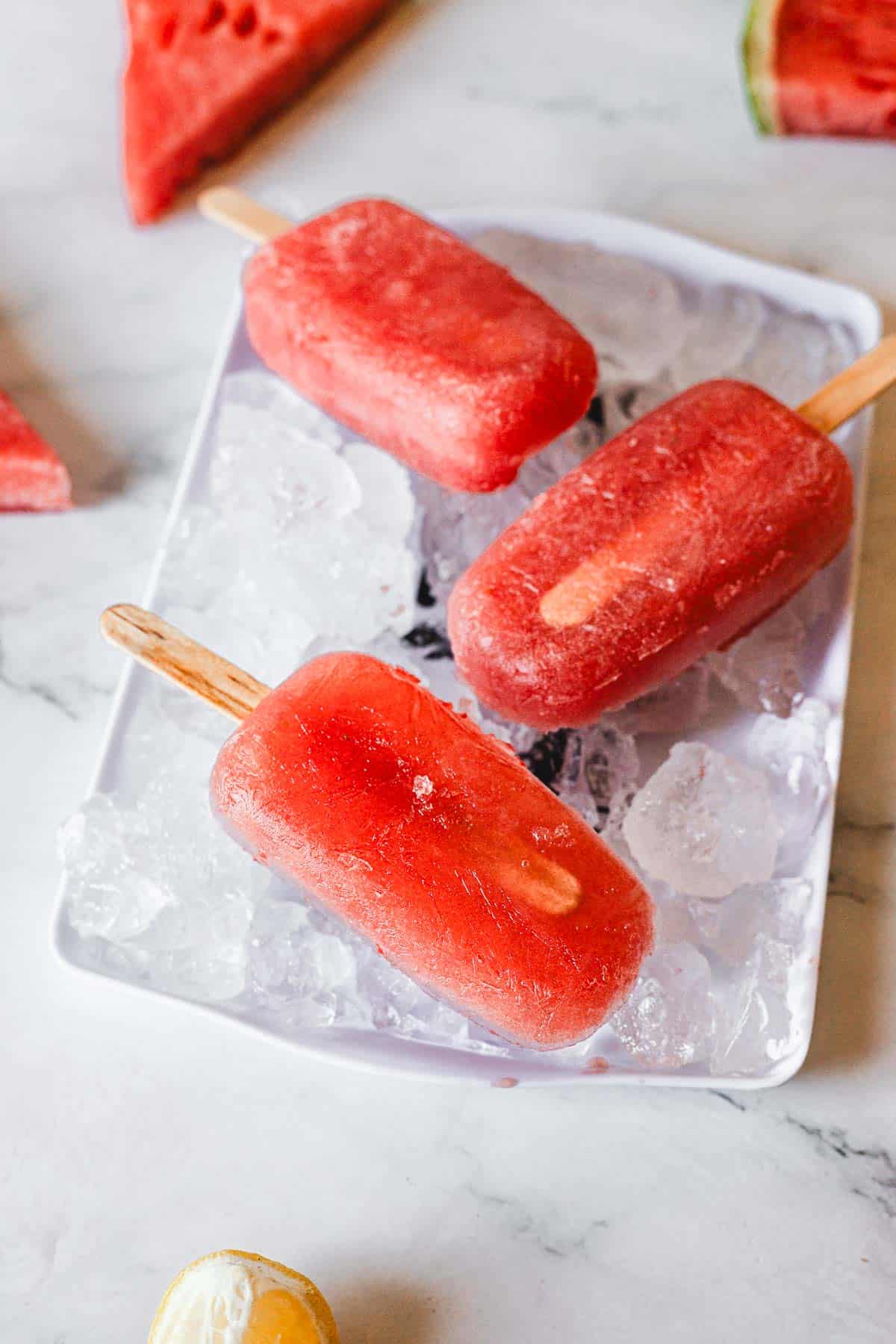 Three red Watermelon Popsicles on wooden sticks rest on a bed of ice in a white dish. Slices of watermelon and part of a lemon are visible around the plate on a light-colored surface.
