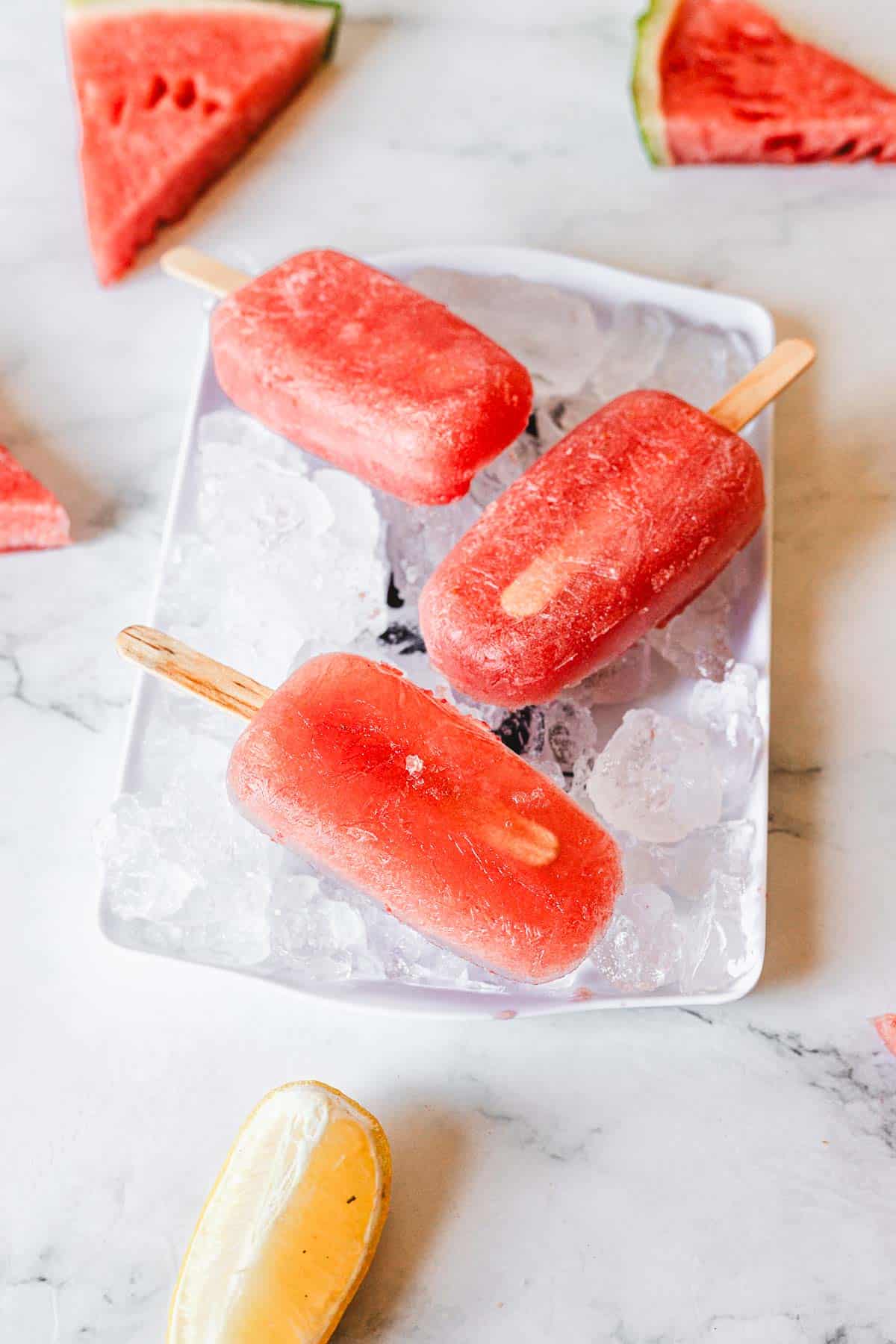 Three red watermelon popsicles on wooden sticks rest on a bed of ice in a white tray, with fresh watermelon slices and a yellow popsicle nearby on a marble surface.