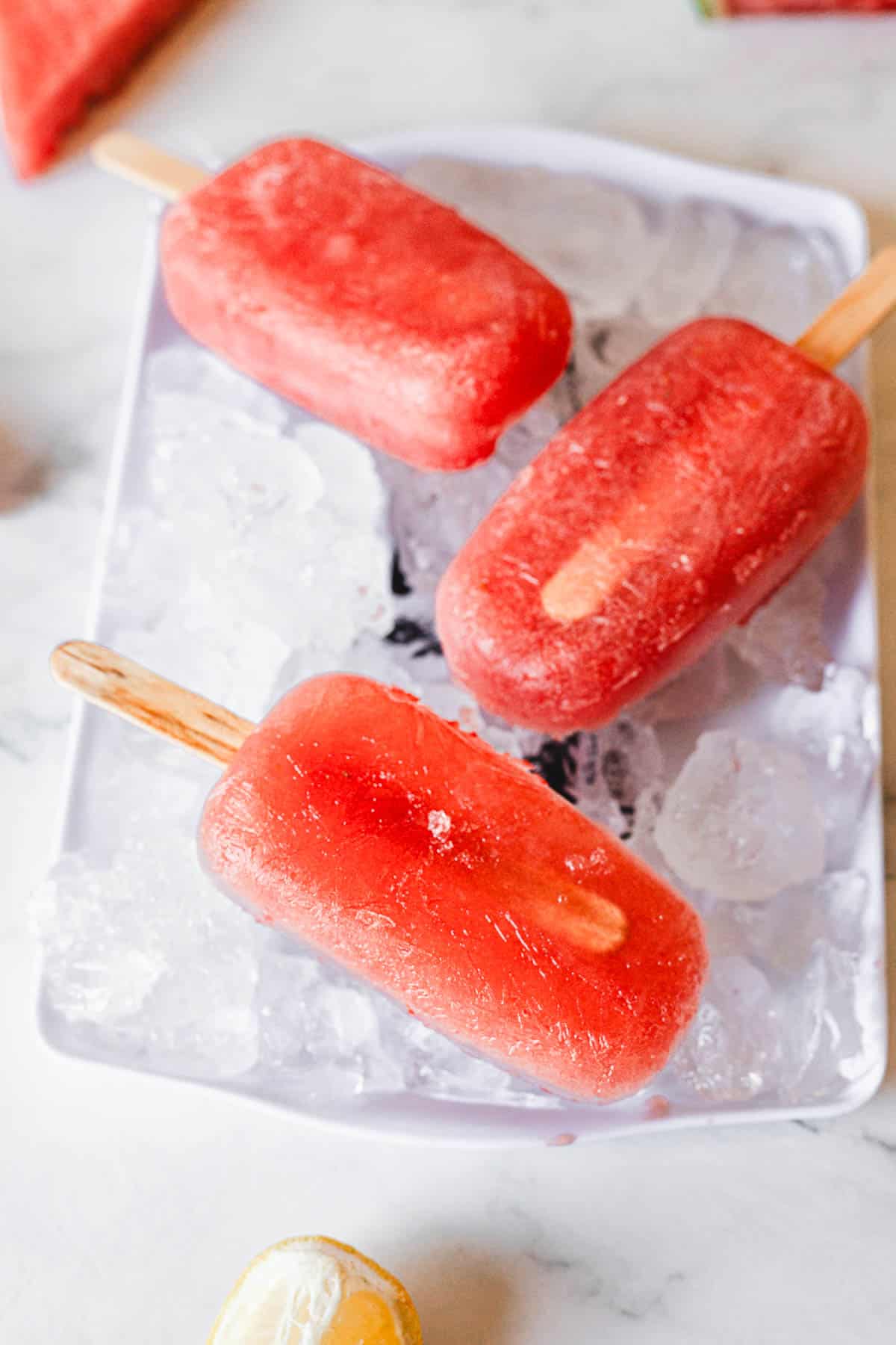 Three red watermelon popsicles are placed on a bed of ice cubes on a white rectangular tray. The background features a marble surface and a partial lemon slice at the bottom of the image.