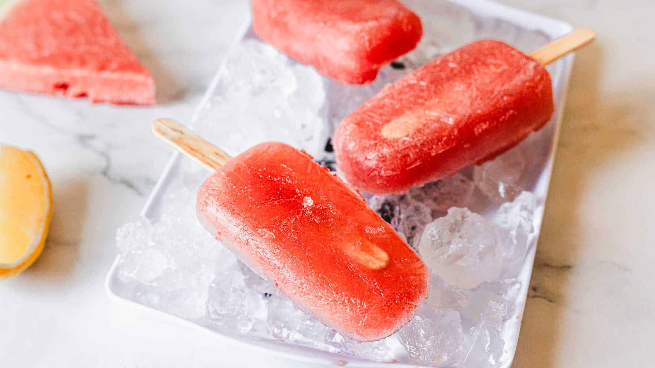 Three red watermelon popsicles are placed on a white tray filled with ice cubes. A slice of watermelon and part of a yellow fruit are visible in the corners of the image on a light surface.