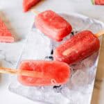 Three red Watermelon Popsicles on wooden sticks rest on a tray filled with ice cubes. Pieces of watermelon are scattered around the tray on a light-colored surface.