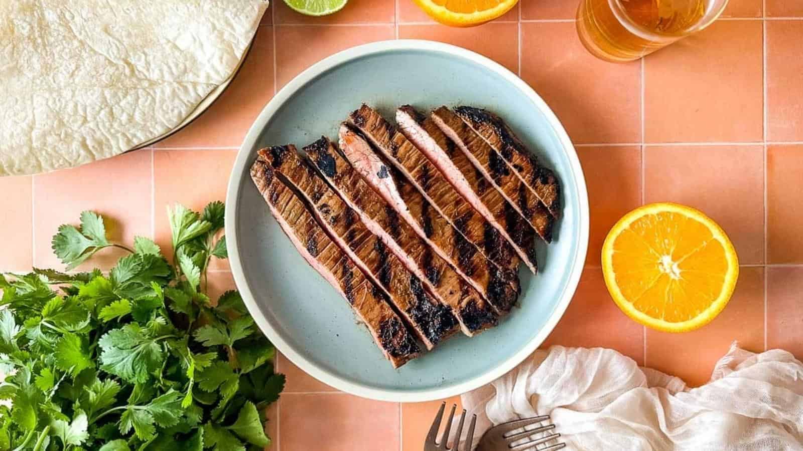 Sliced carne asada sits on a light blue plate surrounded by sliced oranges, limes, a bunch of cilantro, tortillas, and a glass of beer.