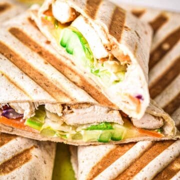 Two grilled wraps are cut in half and stacked, showing fillings of sliced chicken, cucumber, lettuce, and other vegetables inside toasted tortilla bread with grill marks.